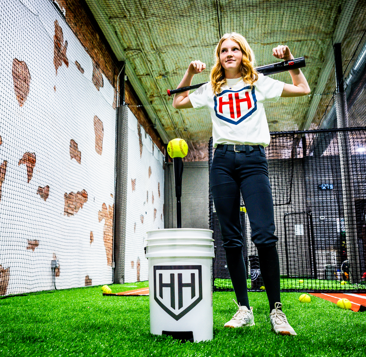Teen softball player holding bat over shoulders and standing next to Home Run Hero ball bucket and tee