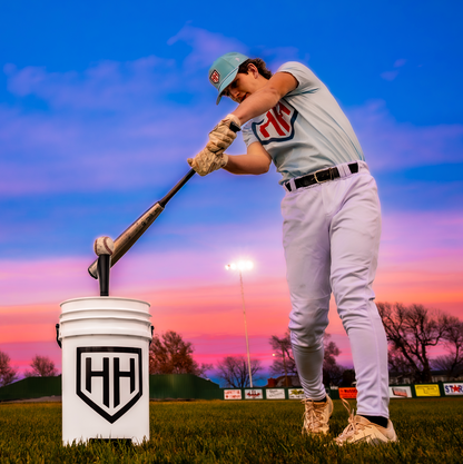 Baseball player hitting baseball off of Home Run Hero tee with sunset in background