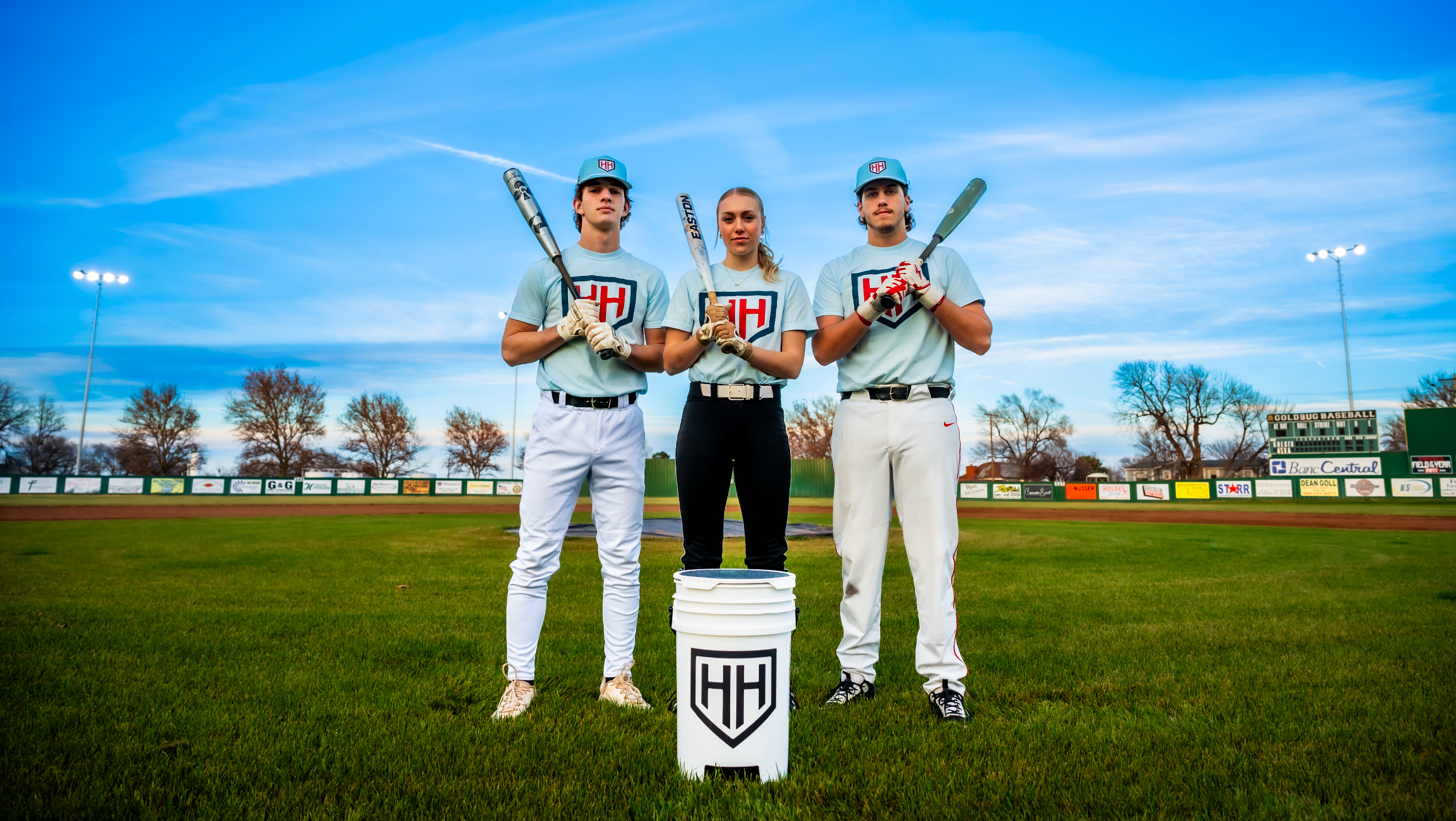 Three baseball and softball players standing in field holding bats with Home Run Hero bucket in front of them