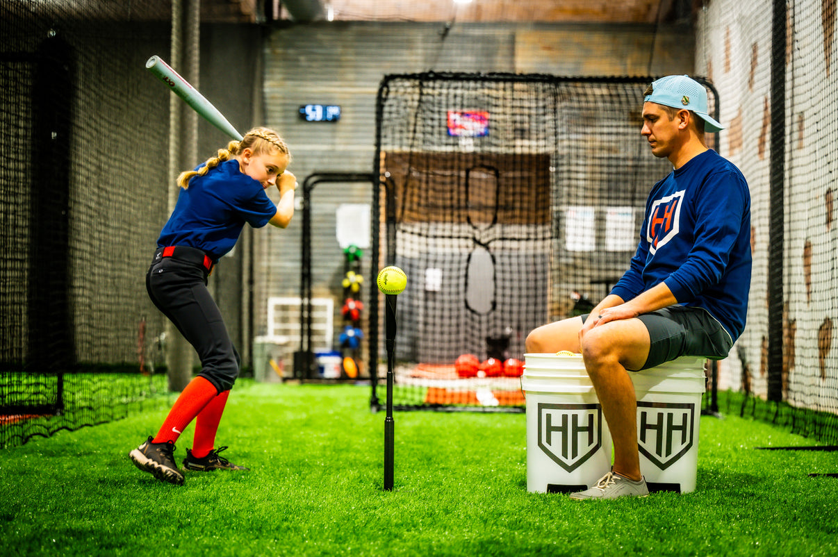 Young softball player hitting softball off of Home Run Hero tee with coach sitting on ball bucket