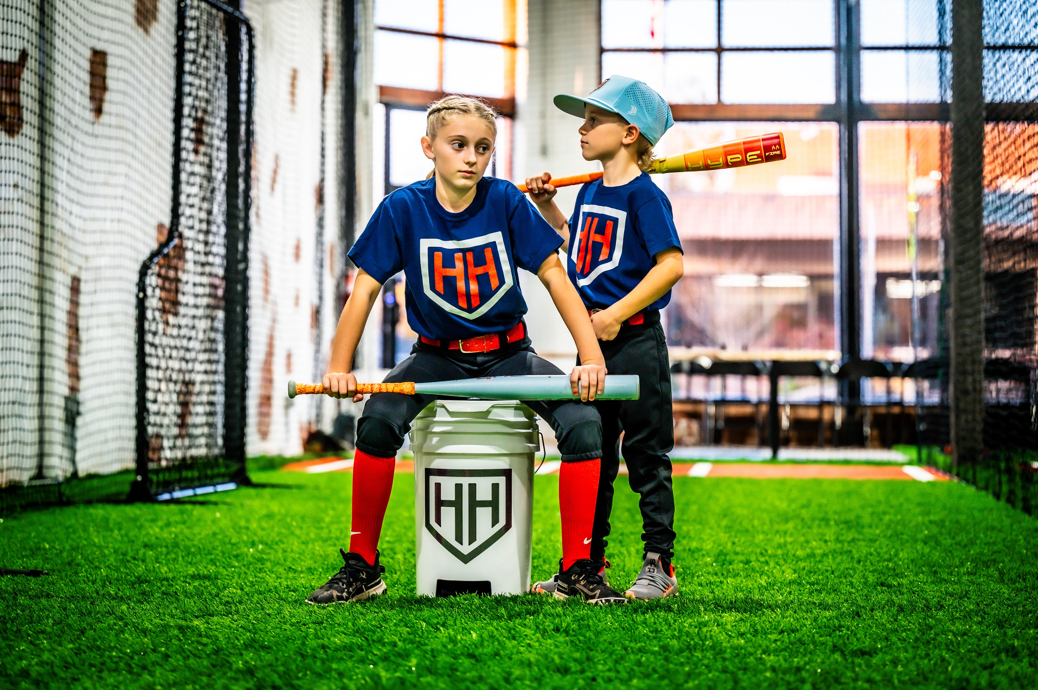 Young softball player sitting on Home Run Hero ball bucket with young baseball player standing behind